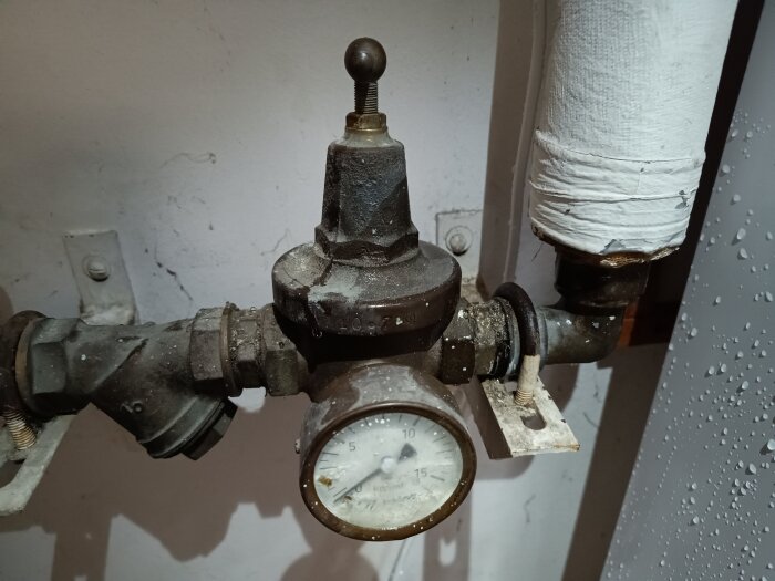 Industriell mätare, rör, ventiler och isolering mot en betongvägg. Något slitet och åldrat.