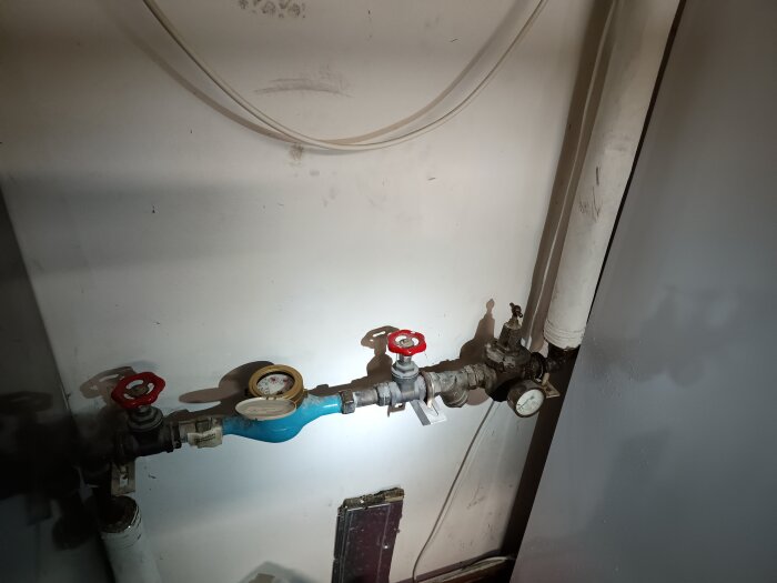 Vattenmätare och ventiler på en vit vägg med synliga rör och diverse anslutningar.