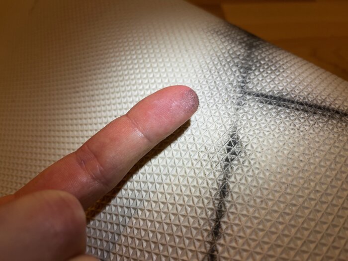 Ett finger vidrör en yta med mönster och en svart ritning eller skada.