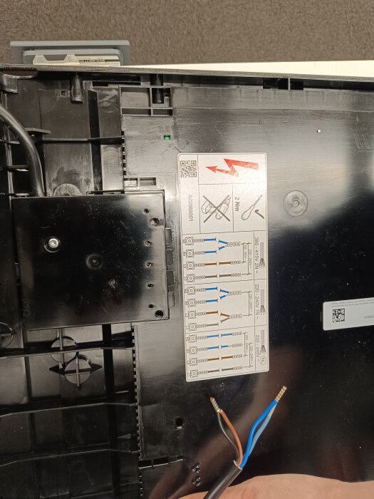 Ett öppet elektriskt appratskal med varningsetikett, ledningar och QR-kod. Verkar vara under underhåll eller reparation.