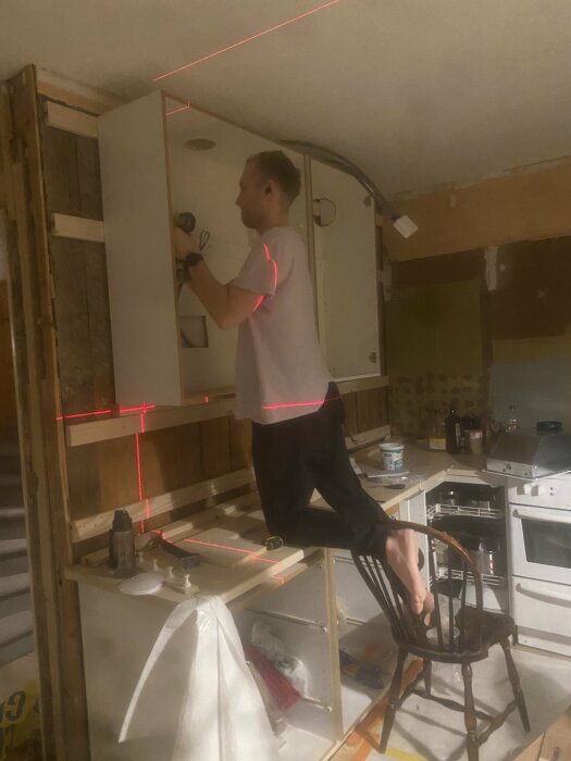Person använder lasernivå vid renovering inomhus, står på stol, kök i bakgrunden, oavslutade väggar, arbetsmiljö.