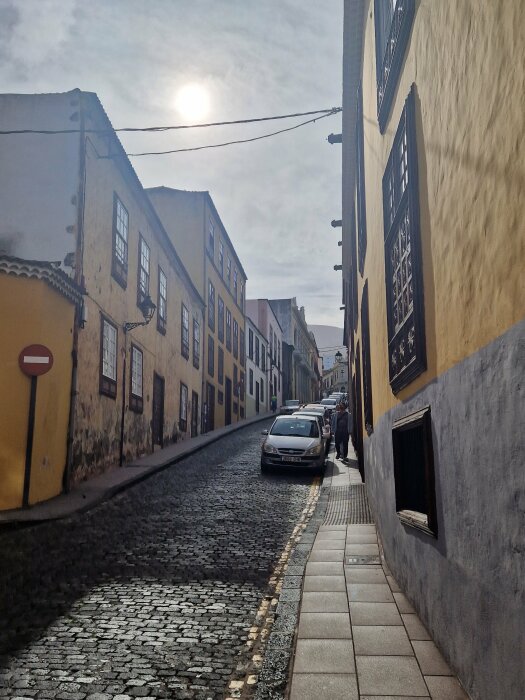 Gammal stadsgata, kullersten, traditionella byggnader, parkerade bilar, soligt, människor promenerar, europeisk arkitektur.