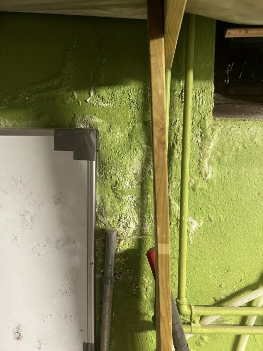 Oorganiserat källarrum med grön vägg, rör, träplanka och vit våtrumsskiva.