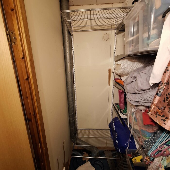 Ett trångt förvaringsutrymme med hyllor, kläder, lådor och en synlig ventilationsrör.