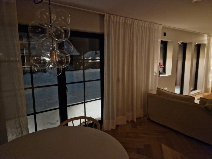 Ett modernt vardagsrum på kvällen med utsikt över snöigt landskap genom fönstret, inredning i neutrala toner.
