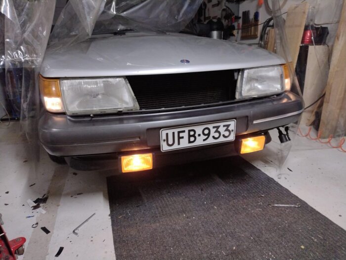 Saab i garage, tända strålkastare och blinkers, verktyg och skyddsplast, nummerskylt synlig.