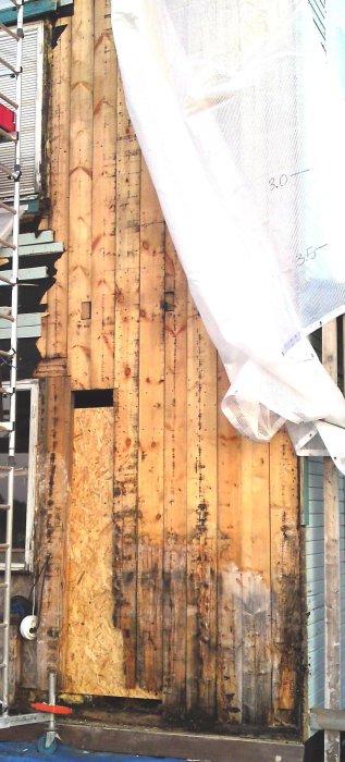 Exterior view of a rotting wooden wall with some boards missing, partially covered by a white tarp. A scaffold and OSB board are visible beside it.