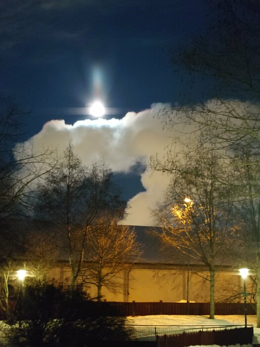 Nattbild, måne bakom moln, ljusstrålar, träd silhuetter, snö, stadsbelysning, industrianläggning, vinteratmosfär.