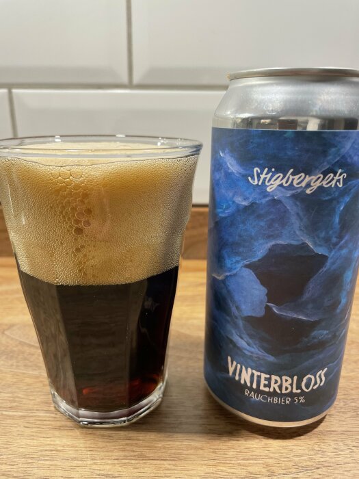 Ölglas med mörkt skum, ölburk med texten "Stigbergets Vinterblomst Rauchbier 5%" på träyta.