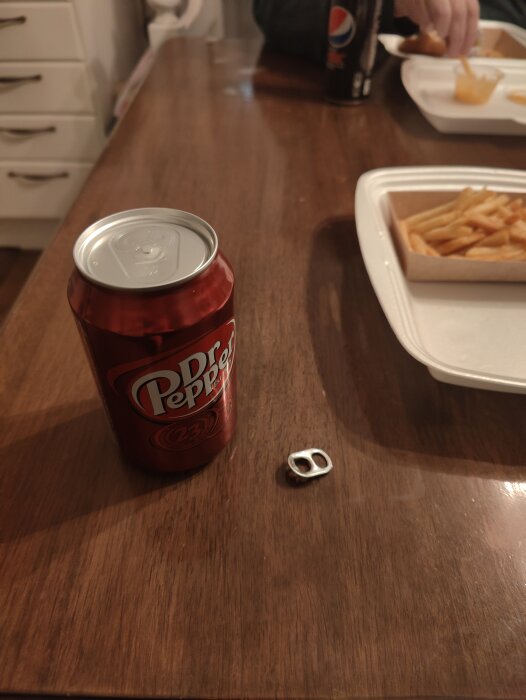 Oöppnad Dr Pepper-burk, ring pull-tab bredvid, pommes frites, suddig person äter, inomhus.