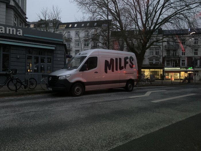 En parkerad vit skåpbil med texten "MILES" vid gatukorsning i skymningen; byggnader och cyklar i bakgrunden.