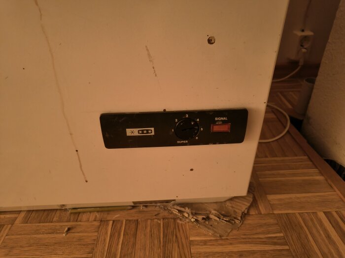 Gammal, sliten, vit apparat med reglagepanel, märken på ytan, vid trägolv och vägg.