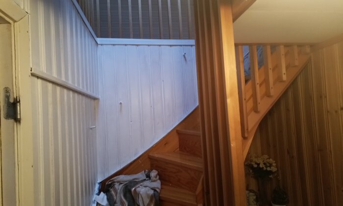 Inomhusbild av en trätrappa med panelvägg, geländer och saker vid foten.