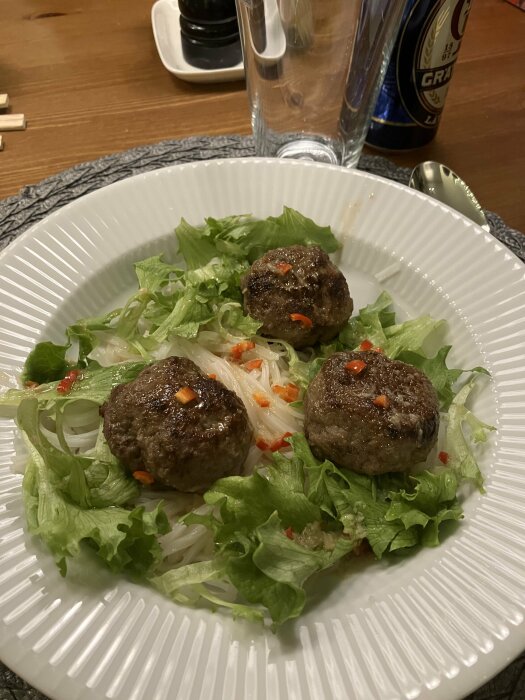 En tallrik med fyra köttbullar, sallad, nudlar, kryddor och en dryckesburk i bakgrunden.