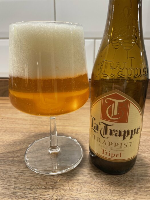 Ett glas med öl och tjockt skum bredvid en La Trappe Trappist Tripel-flaska på bord.