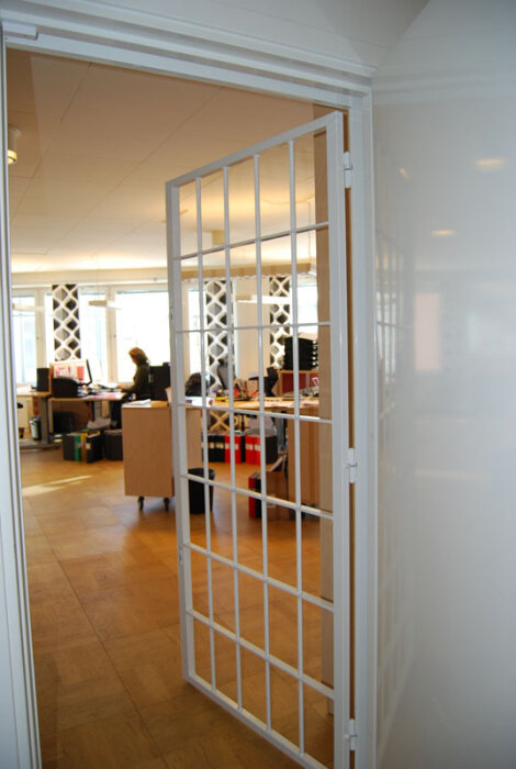 Vit grinddörr öppen till kontorslandskap med arbetsstationer och person i bakgrunden.