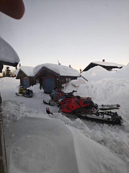 Snöskotrar framför snötäckta stugor i vinterlandskap, klart väder, dagtid.