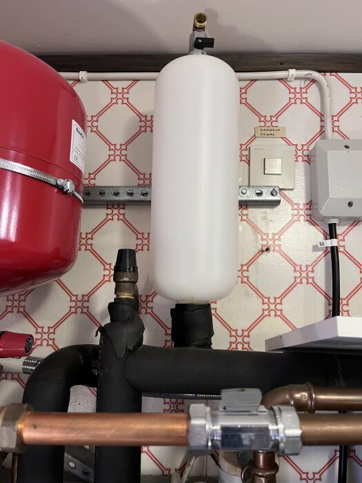 Installation av VVS med expantionskärl, rör, kopplingar och tryckmätare på vägg med mönstrad tapet.