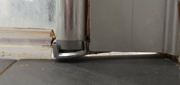Bild av skadat dörrstopp vid golv, nedsmutsad vägg, rostfritt stål, slitage, behov av underhåll/reparation.