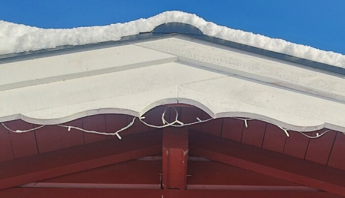 Ett snötäckt tak med vit och röd fasad samt hängande julgransljus under en klarblå himmel.