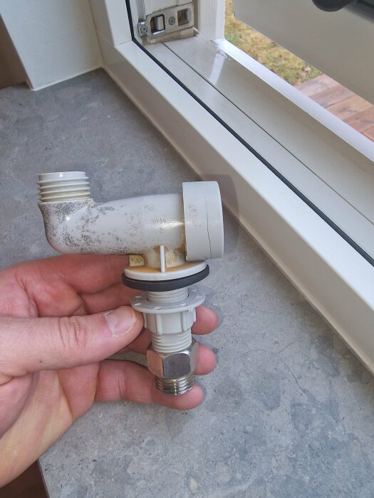 Hand håller plast och metall avloppsdel nära fönster; möjlig reparation eller installation inomhus.