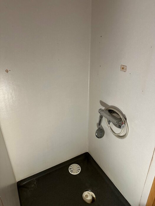 Tomt duschutrymme med handdusch, slitet svart golv, vita väggar, avloppshål, utan monterad duschdraperistång.