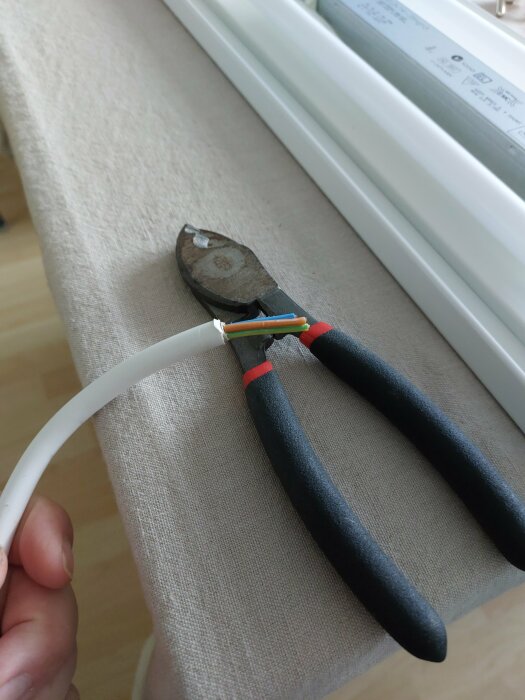 En avbitartång klipper igenom en vit elektrisk kabel med exponerade färgade trådar på en soffiarmstöd.