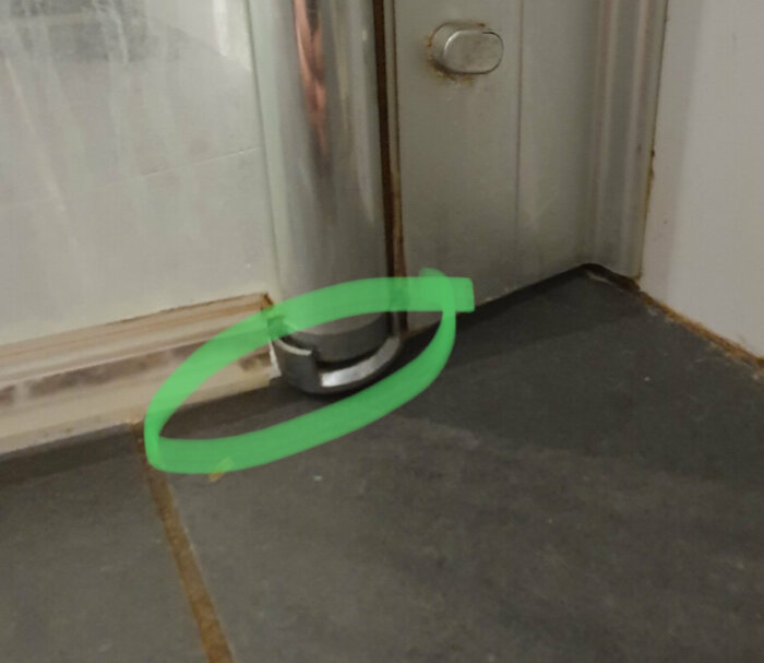 Dörrstopp i metall på ett grått golv, markerat med grön cirkel, nära en dörr.