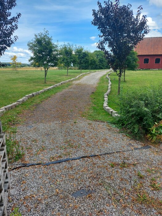 Grusgång, stenkanter, grönt gräs, träd, röd ladugård, blå himmel, moln, lantlig miljö.