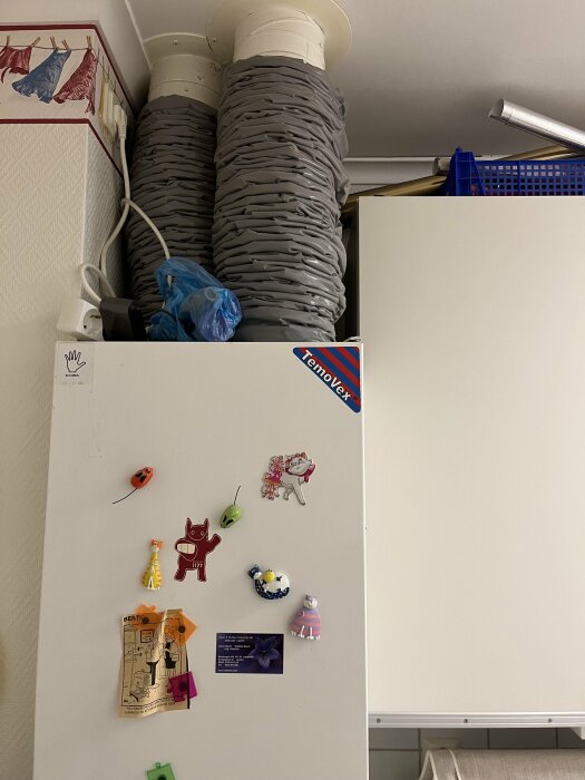 Kylskåp med magneter, färgglada figurer, ventilationsrör, rörig hylla, plastpåsar, hemmiljö.