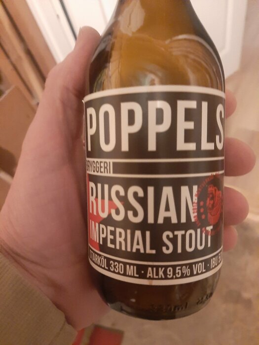 Hand håller Poppels Russian Imperial Stout ölflaska, 330 ml, 9,5% volymandel alkohol, bakgrund oklar.