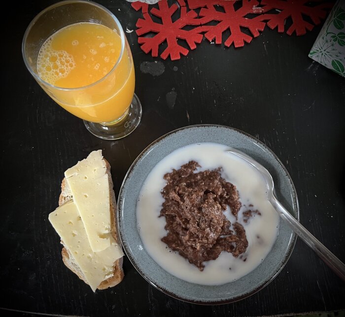 Frukost med havregrynsgröt, mjölk, ostsmörgås och apelsinjuice på ett mörkt bord med röda snöflingedekorationer.