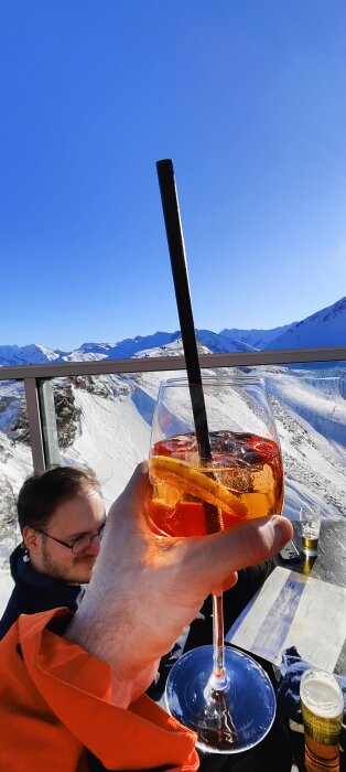 Drink i fokus, man vid bord, snöklädda berg, soligt, utomhus, avkoppling, terrass, vintersemester.