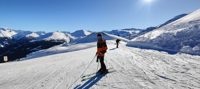 Skidåkare på snöklädd bergssluttning, under klar himmel, imponerande bergslandskap i bakgrunden.