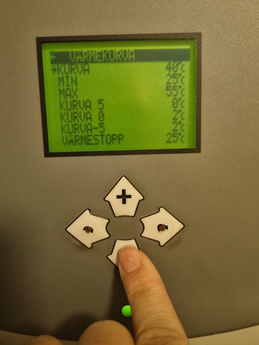 En hand interagerar med en kontrollpanel med LCD-skärm och knappar. Text på skärmen antyder temperaturinställningar på svenska.