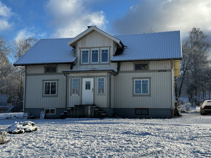 Tvåvånings trähus med snötäckt tak och mark, soligt vinterväder, bil parkerad till höger.