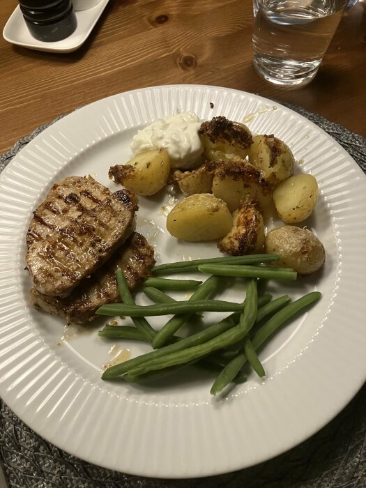 Grillad köttbit, rostade potatisar, gröna bönor och vit sås på en tallrik. Glas vatten. Trämöbler.