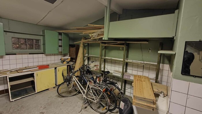 Oorganiserad källare med cyklar, skräp, skåp och byggmaterial. Slitna, omatchande möbler och sjaskiga väggar.