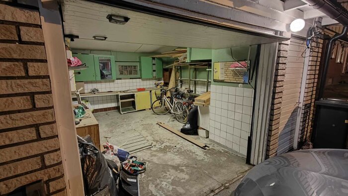 Öppen garageverkstad med cyklar, verktyg, skräp, och hyllor med diverse föremål. Oorganiserat och används för förvaring.