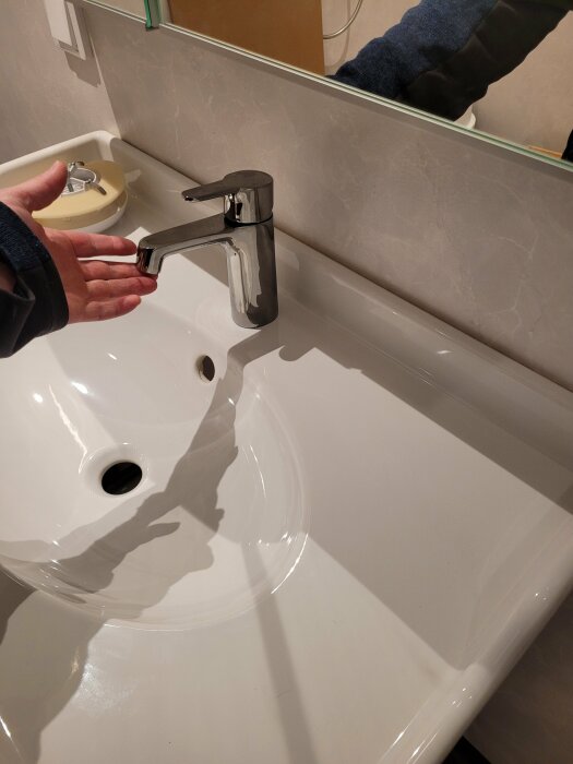 Person testar vattenkranen över en vit handfat; ingen vattenflöde; spegel och kakelvägg syns i bakgrunden.