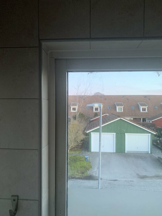 Utsikt från ett fönster visar garage, träd och byggnader vid skymning eller gryning. Smuts på glaset.