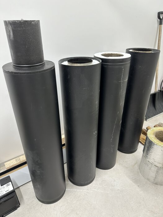 Fyra stora svarta rör står på golv, avtagbara toppar, industriell miljö, vit vägg bakgrund.