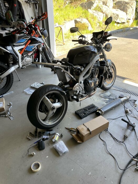 Motorcykel utan bakhjul i garage, verktyg och delar spridda på golvet, annan motorcykel i bakgrunden.