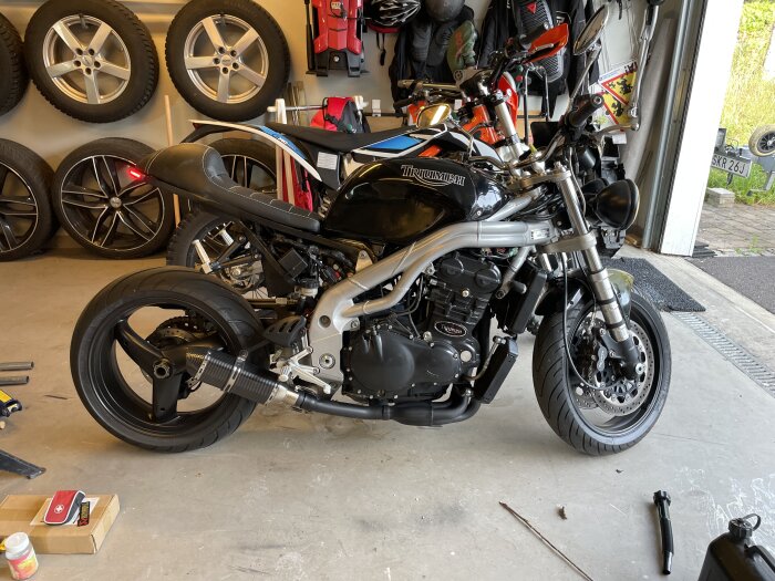 Motorcykel Triumph i garage med däck och verktyg runt omkring. Ounderhållsstil med öppen dörr.