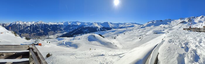 Ett panoramafoto av ett snötäckt bergslandskap med skidbackar och klarblå himmel.