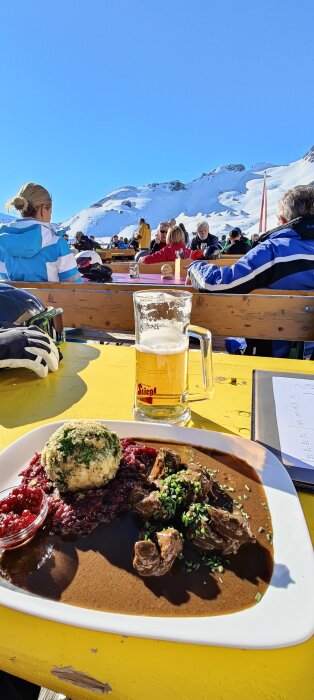 Solig bergsterrass med öl och mat, människor i vinterkläder, avkopplande efter skidåkning, blå himmel och snötäckta toppar.