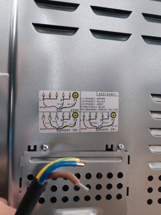 En hand håller en avskalad elkabel framför en kopplingsschemaetikett på en metallisk enhet.