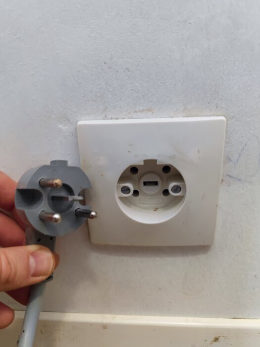 Hand håller europeisk stickkontakt nära oanvänd vägguttag, smutsiga märken runtom, grå enhet och kabel.