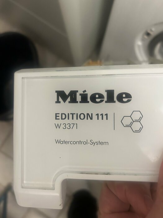 Vit plastdetalj med logotyp "Miele", text "EDITION 111 W3371", "Watercontrol-System". Hand håller taget.