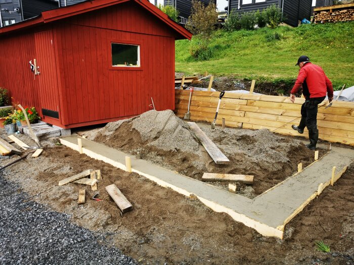 Person arbetar med grundläggning utomhus, sand, plankor, verktyg, röd stuga, grått molnigt väder.